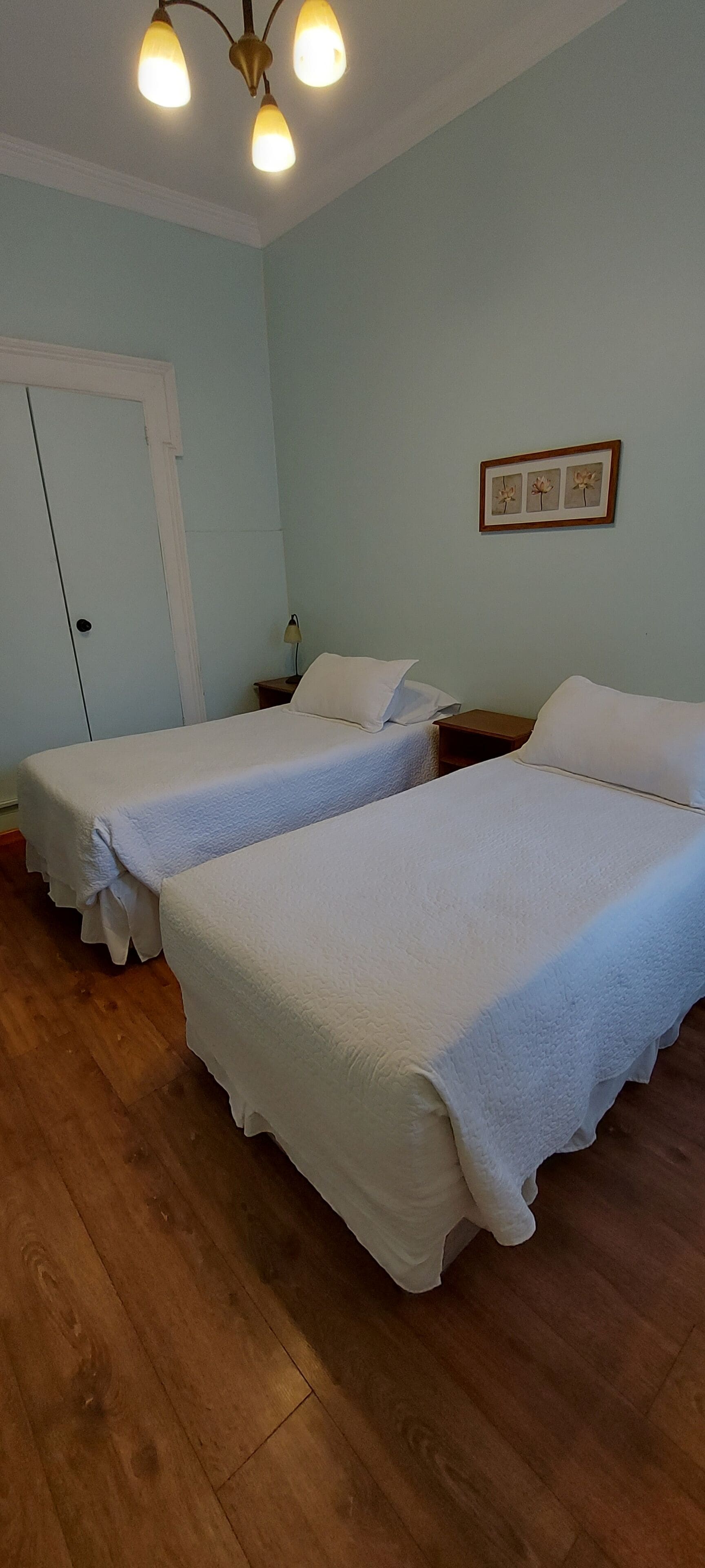 A room with two beds, wooden flooring, a white door, and a ceiling light fixture.