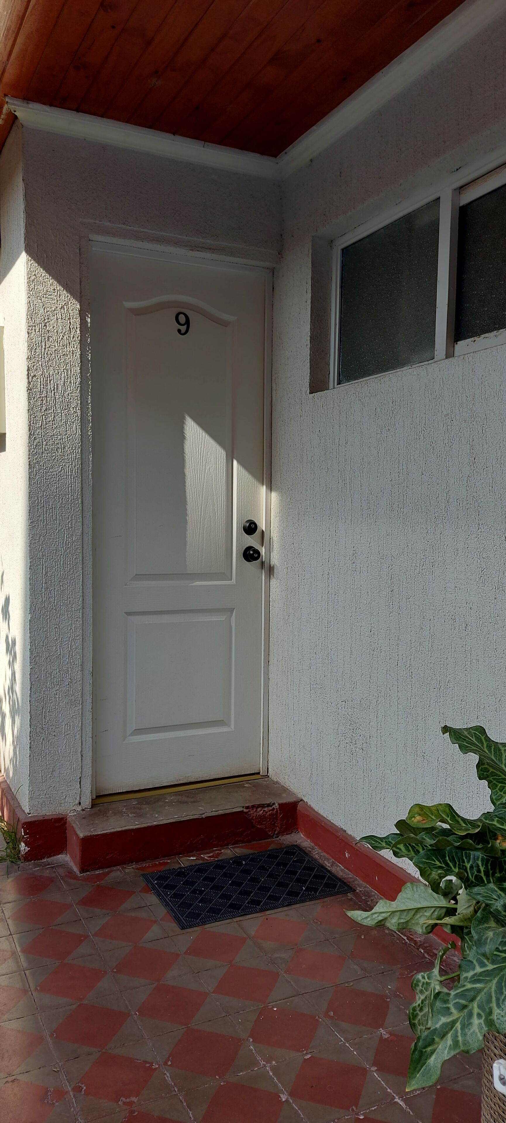 Une porte blanche portant le numéro 9, une petite fenêtre au-dessus, et un porche carrelé de rouge et de beige avec un paillasson noir.