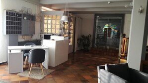 A modern kitchen with white cabinets, a dining area with a black chair, and a large window with a view of a colorful building. - Le Malatras (Tullins)