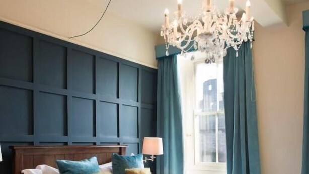A bedroom with a large bed, a chandelier, and a view of the city through a window. - The Priory (Leyburn)
