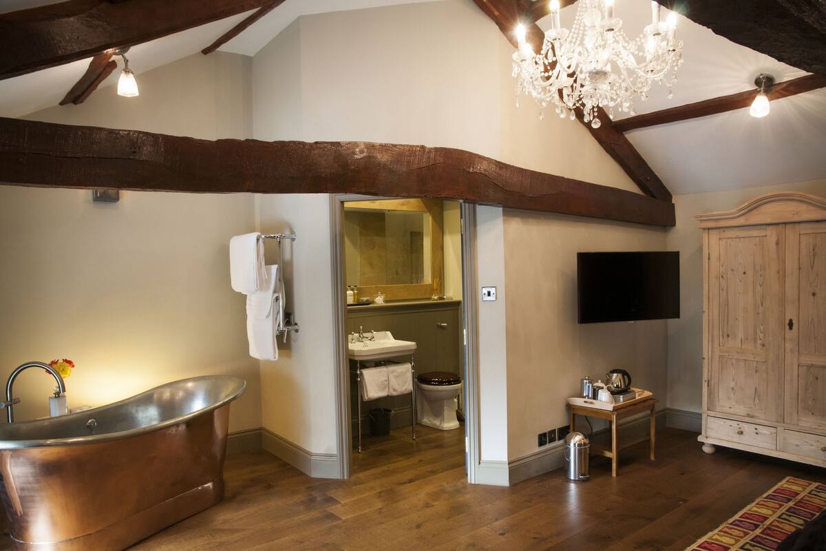 A rustic bathroom with a copper bathtub, wooden beams, a chandelier, and a wooden cabinet.