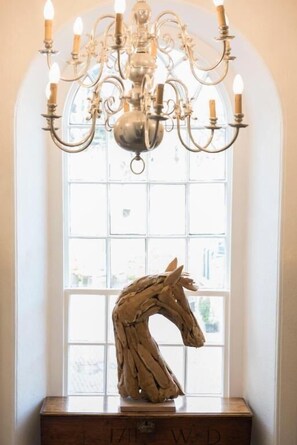 A chandelier with multiple arms and candle-like lights, a wooden horse sculpture, and a window with a view of buildings. - The Priory (Leyburn)