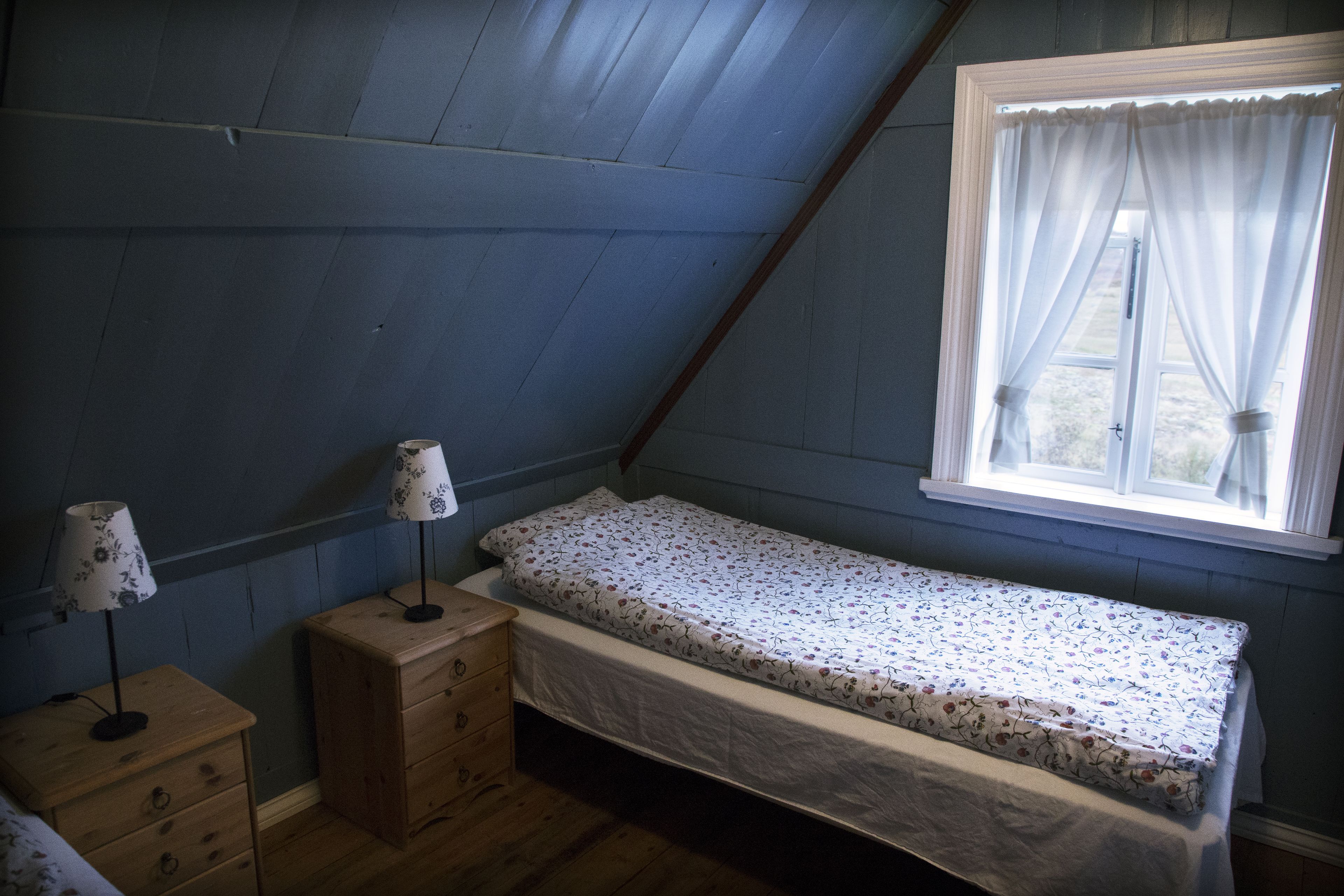 Une petite chambre avec un lit simple, un plafond en pente, une fenêtre avec des rideaux blancs, une commode en bois avec des tiroirs et une lampe de chevet.