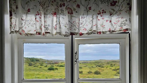 Ein Fenster mit floralem Vorhang und Blick auf ein grünes Feld.