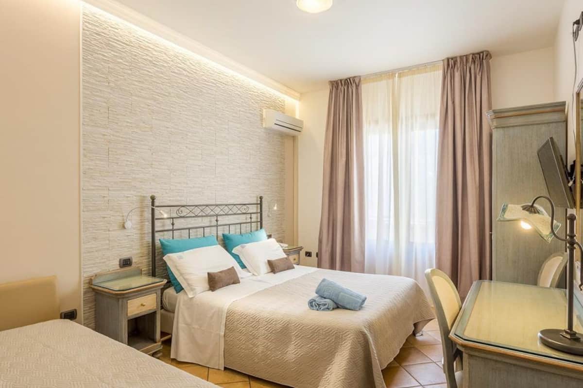 A hotel room with a bed, bedside table, lamp, and a view of the outside through curtains.