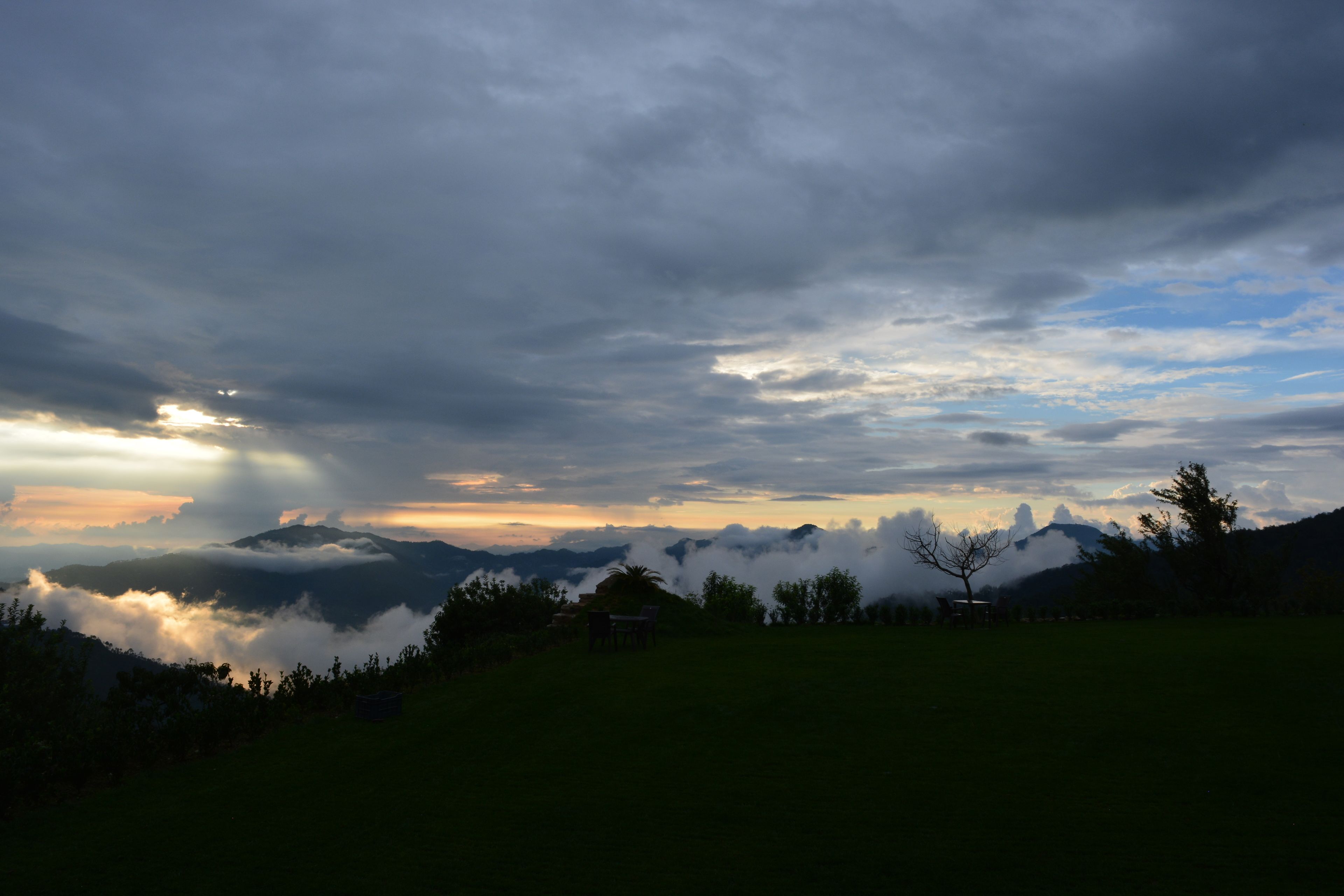 A landscape with a chair and table in a grassy field, overlooking a mountain range with clouds.