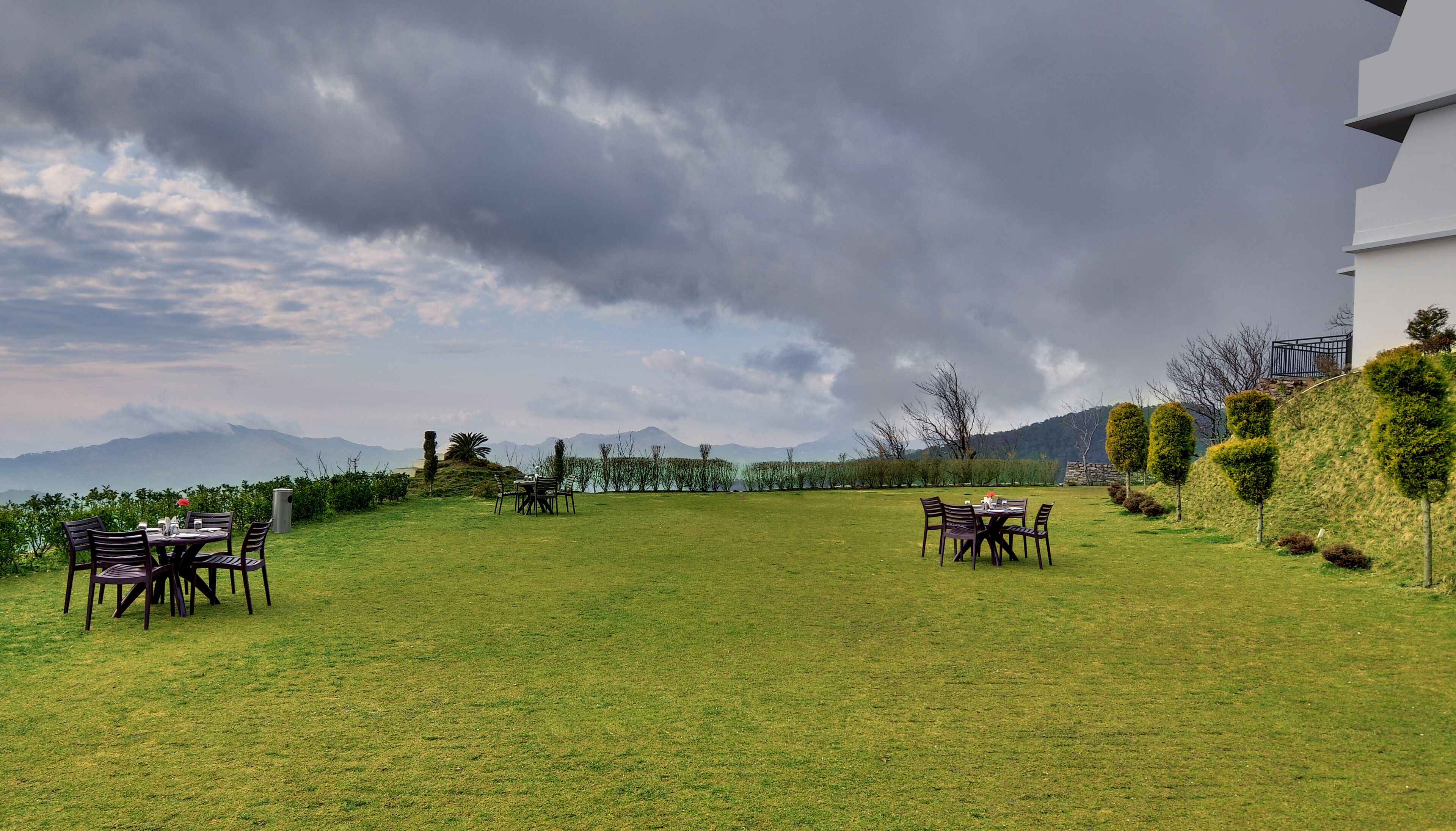 A grassy outdoor area with two dining tables and chairs, surrounded by neatly trimmed hedges and a view of distant mountains under a cloudy sky.