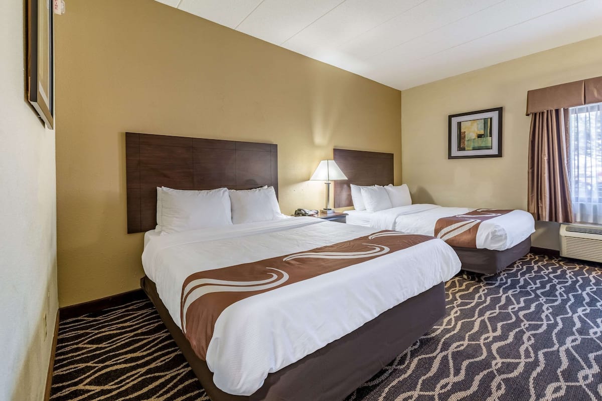 A hotel room with two beds, a large window with curtains, a wall-mounted air conditioner, and a framed picture on the wall.