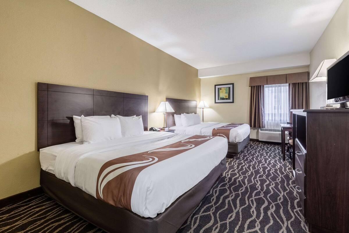 A hotel room with two beds, a flat-screen TV, a desk, and a patterned carpet.