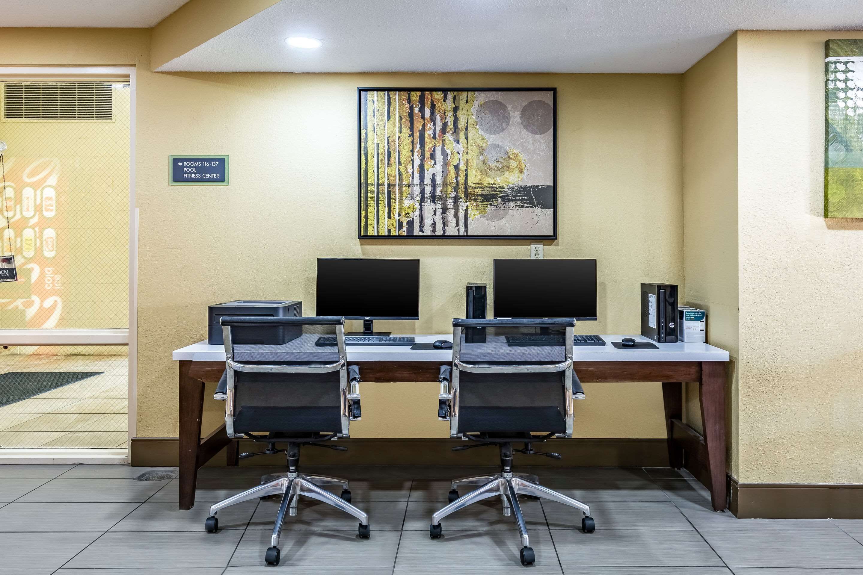 A conference room with two desks, each with a computer monitor, keyboard, and mouse. There are two office chairs, a large abstract painting on the wall, and a sign indicating the room number and purpose.