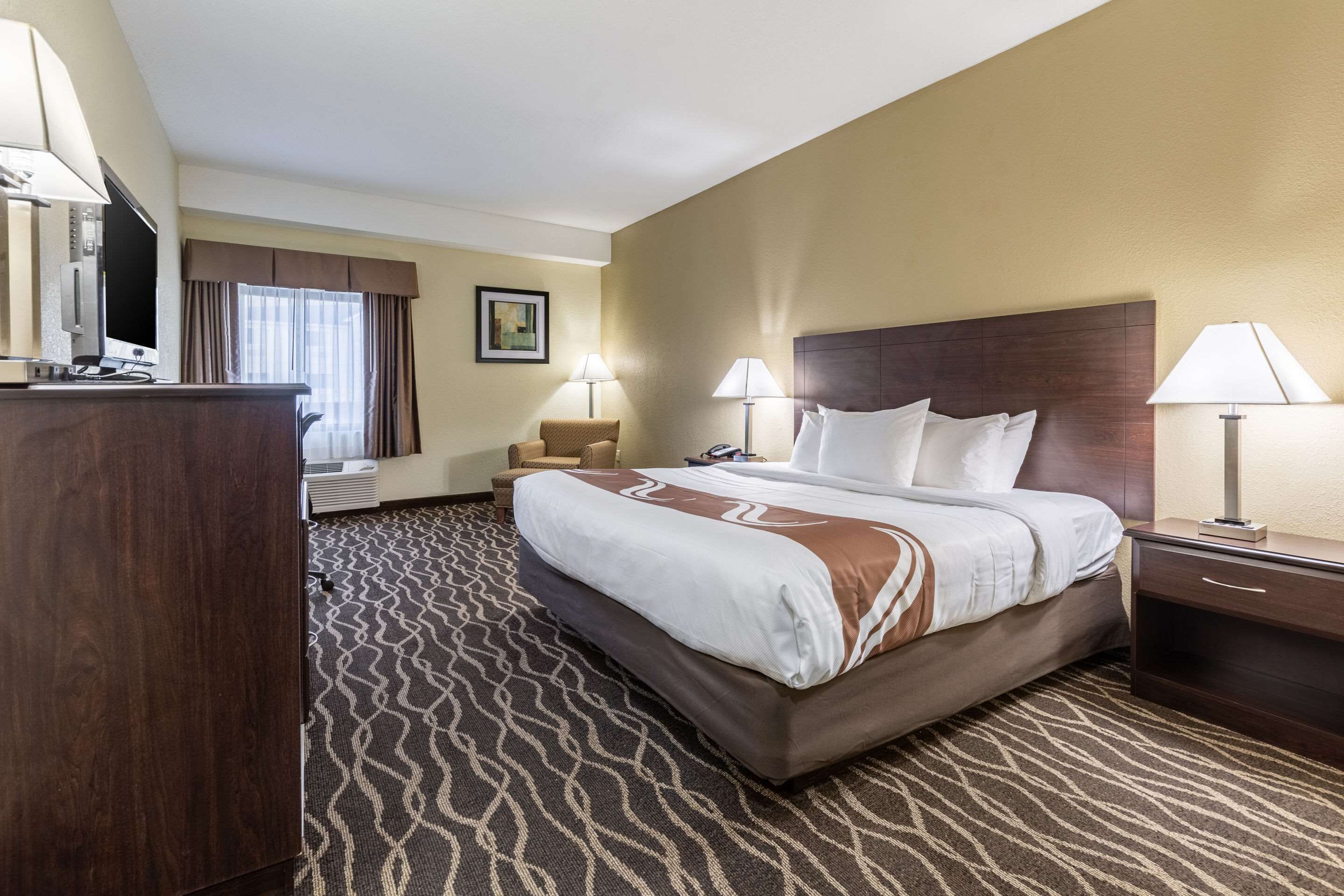 A hotel room with a large bed, two bedside tables, a desk with a lamp, a chair, a TV, and a window with curtains.