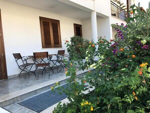 Un área de estar al aire libre con una mesa y sillas, un pequeño jardín con diversas flores, y un edificio con paredes blancas y contraventanas de madera.