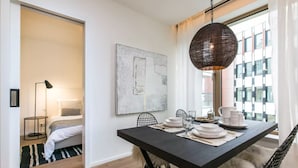 A modern dining area with a dark table set for four, a large abstract painting on the wall, and a view of the city through large windows. - Flawless Apartments Pod Děkankou (Prague)