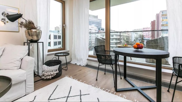 A modern living room with a sofa, a round table with chairs, a small side table, and a large window offering a city view. - Flawless Apartments Pod Děkankou (Prague)