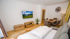 A hotel room with a bed, a sofa, a dining table with chairs, a TV mounted on the wall, and a clock. - Apartment Summer & Winter Fun II (Zell am See)