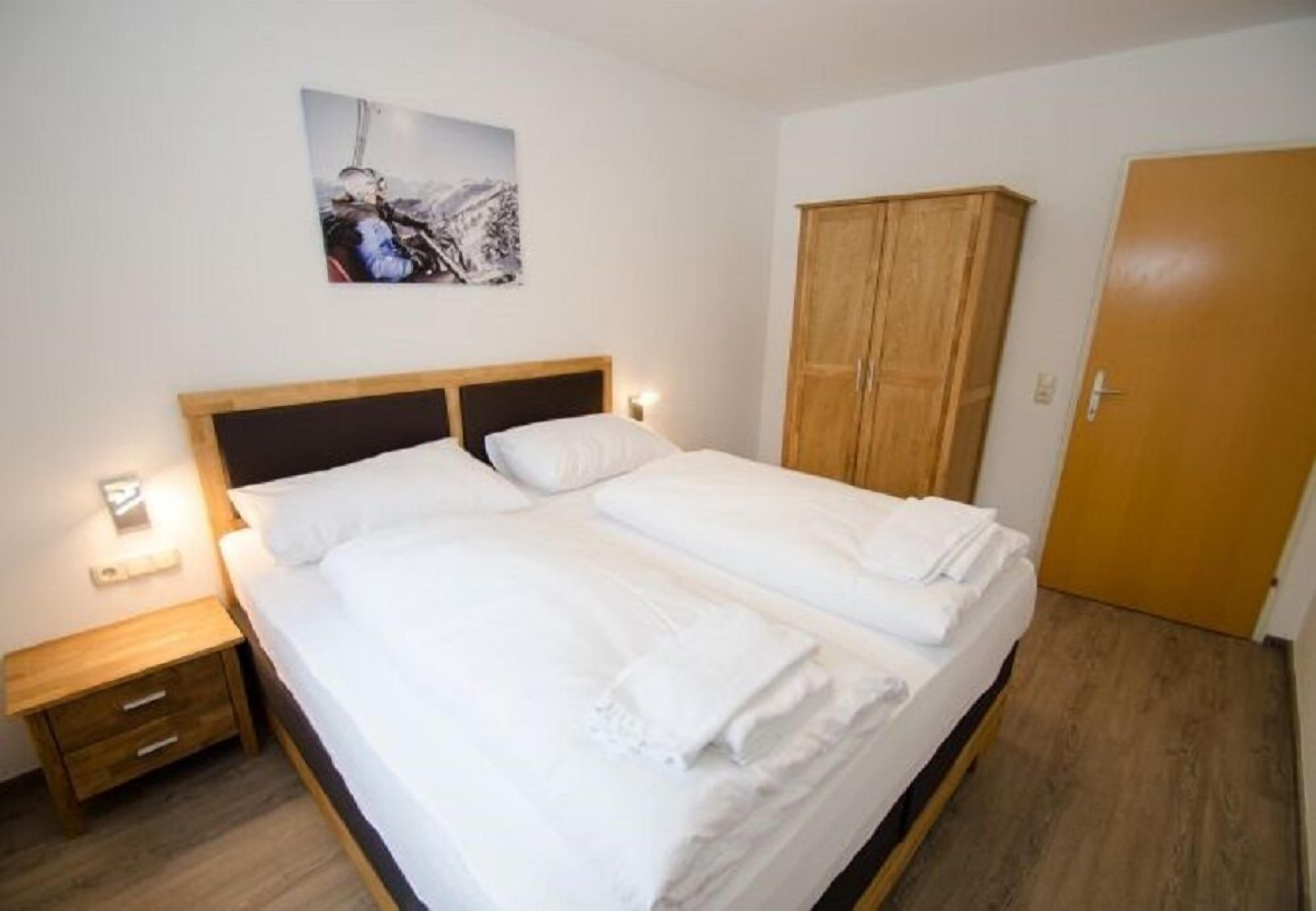 Una cama doble con ropa de cama blanca, un cabecero de madera, una mesita de noche con una lámpara, un armario de madera y una puerta.