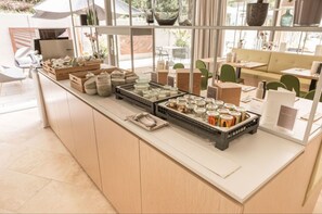 A cafe with a long counter displaying various jars and containers, a seating area with green chairs, and a view of the outdoors. - Das ACKER Hotel (Neuburg)