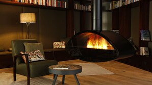 A cozy living room with a modern fireplace, a comfortable sofa, a small side table, and a bookshelf filled with books. - Das ACKER Hotel (Neuburg)