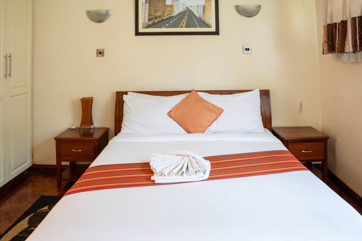 A neatly made bed with a striped bedspread, a folded towel, and a single orange pillow.