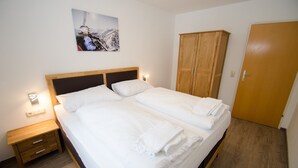 Una cama doble con ropa de cama blanca, cabecera de madera, mesita de noche con cajones, armario de madera y un cuadro en la pared.