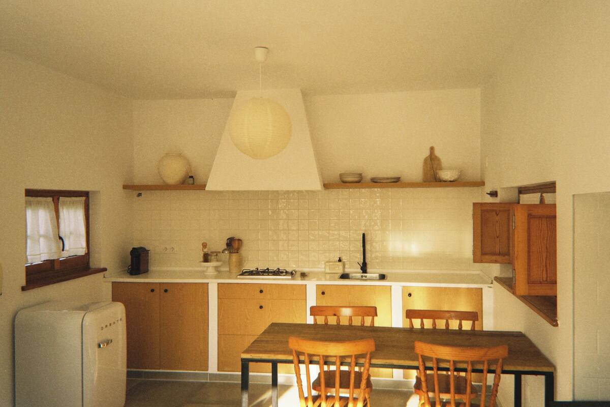 A compact kitchen with wooden cabinets, a dining table with chairs, a hanging light fixture, and a window with curtains.