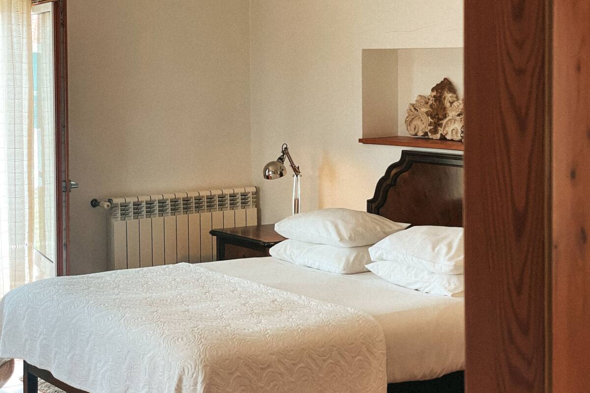A bedroom with a wooden bed, white linens, a bedside table with a lamp, a radiator, and a mirror on the wall.