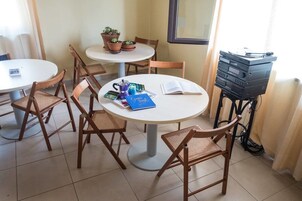 Una habitación con dos mesas redondas, cada una rodeada de sillas de mimbre. Hay plantas en macetas sobre una mesa, una pila de libros y una cafetera en la otra.