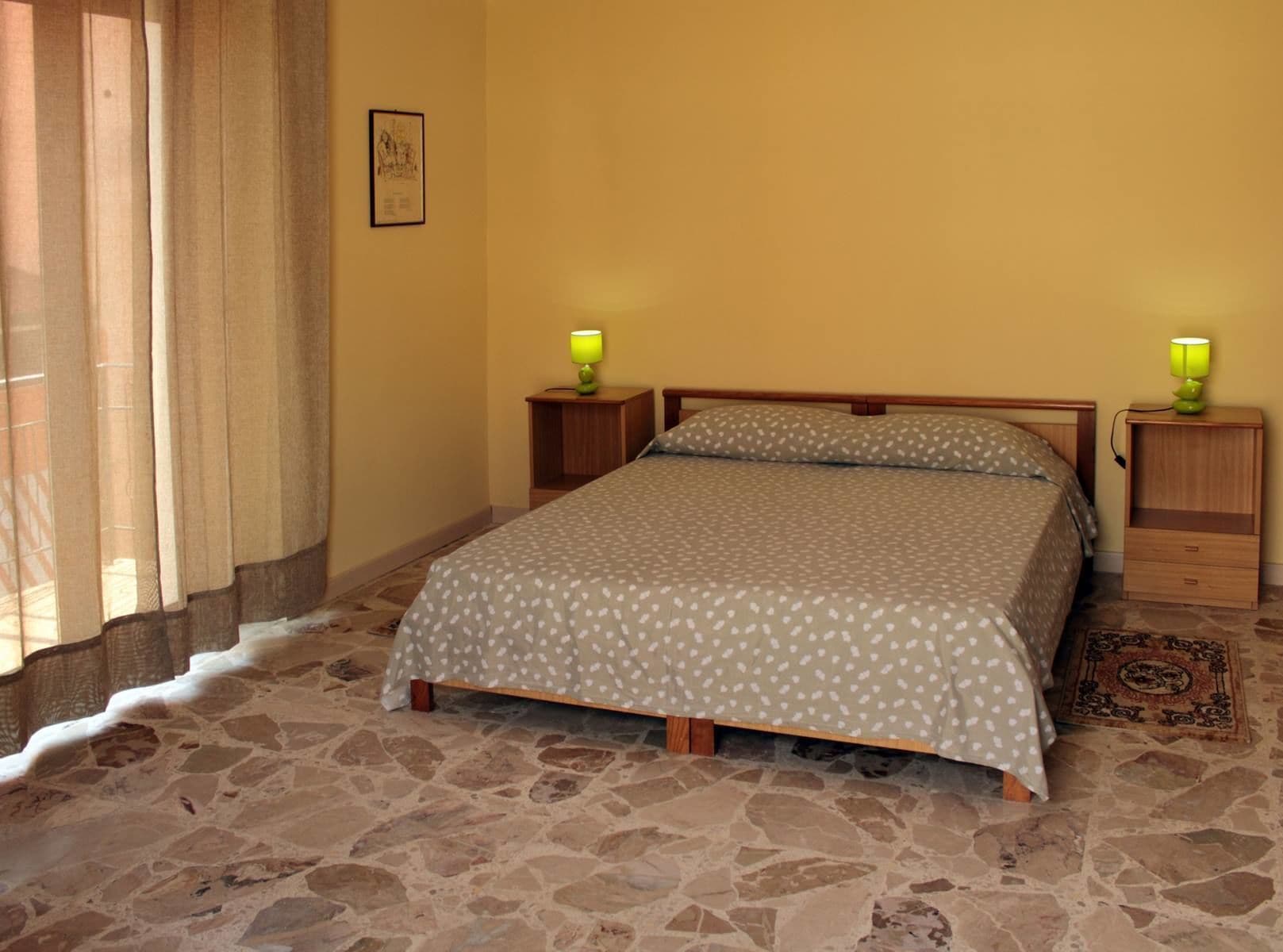 A bedroom with a bed, two nightstands, a lamp, a rug, and a wall with a framed picture.