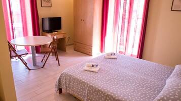Una habitación pequeña y luminosa con una cama, un escritorio con televisor, una silla y una mesa.