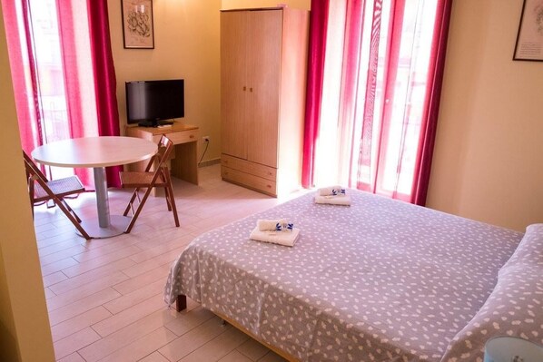 A small, well-lit room with a bed, a desk with a TV, a chair, and a table.