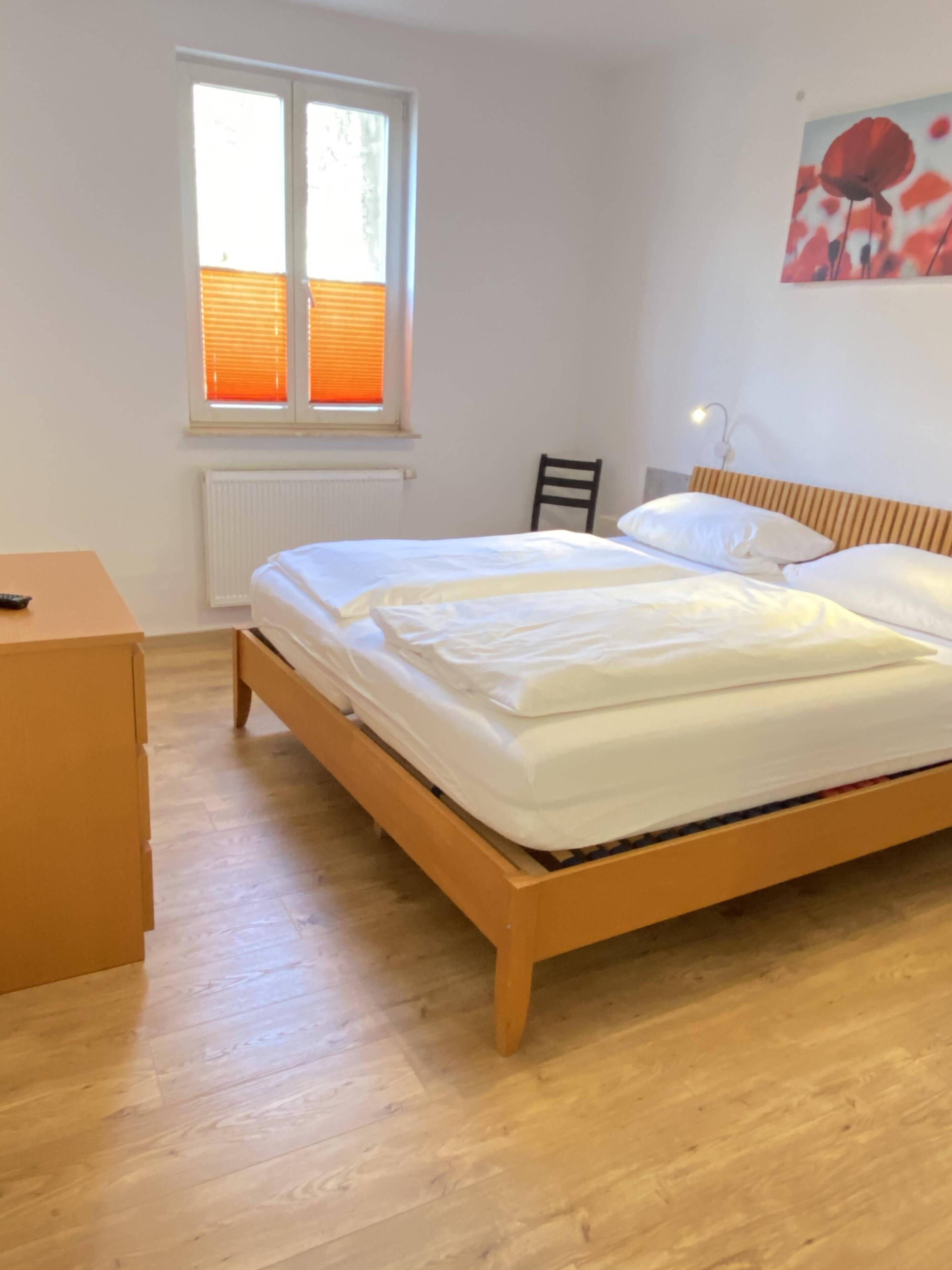 Een slaapkamer met een houten bed, een houten dressoir, een raam met zonwering en een schilderij aan de muur.