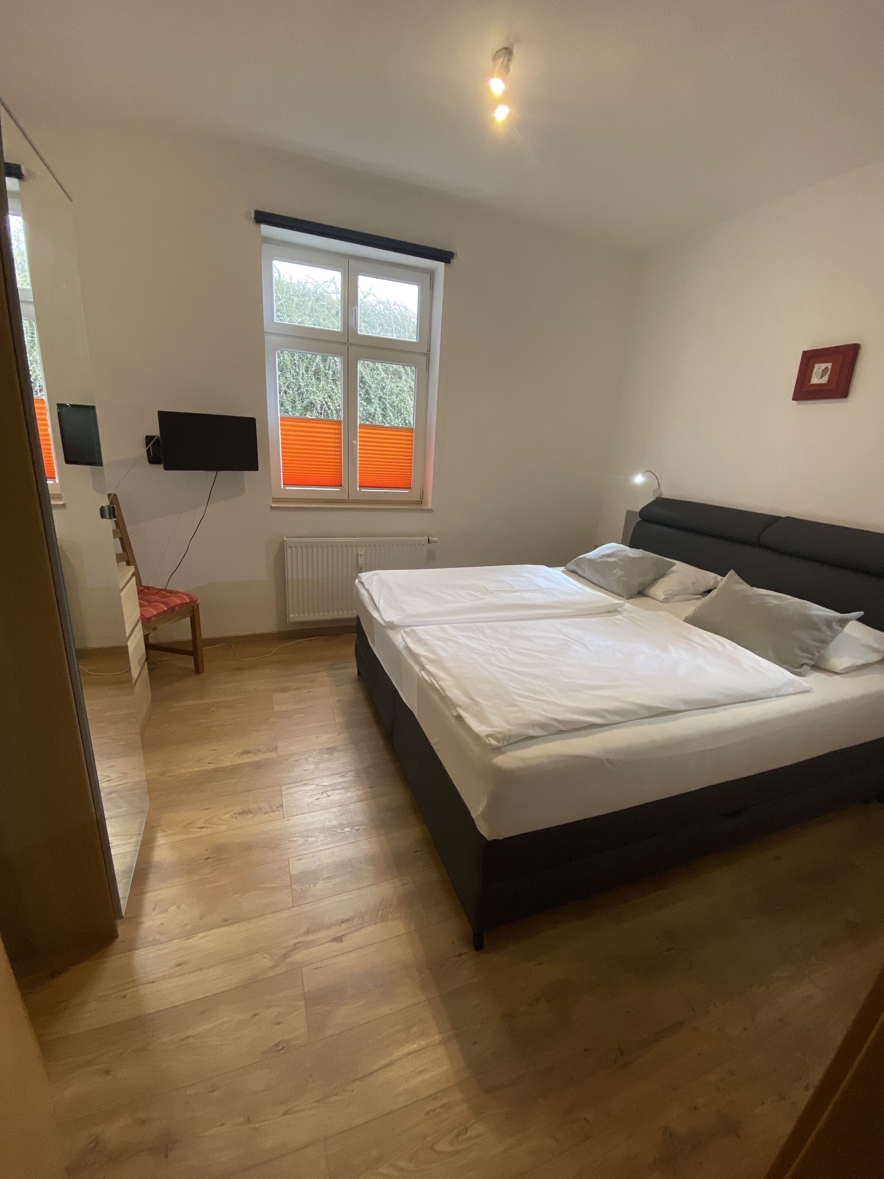 Ein Schlafzimmer mit einem Bett, einem Schreibtisch, einem Stuhl, einem Fenster mit Jalousien und einem am Wand montierten Fernseher.