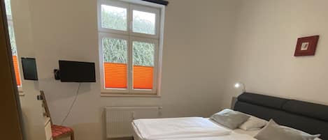 Ein Schlafzimmer mit einem Bett, einem Schreibtisch, einem Stuhl, einem Fenster mit Jalousien und einem am Wand montierten Fernseher.
