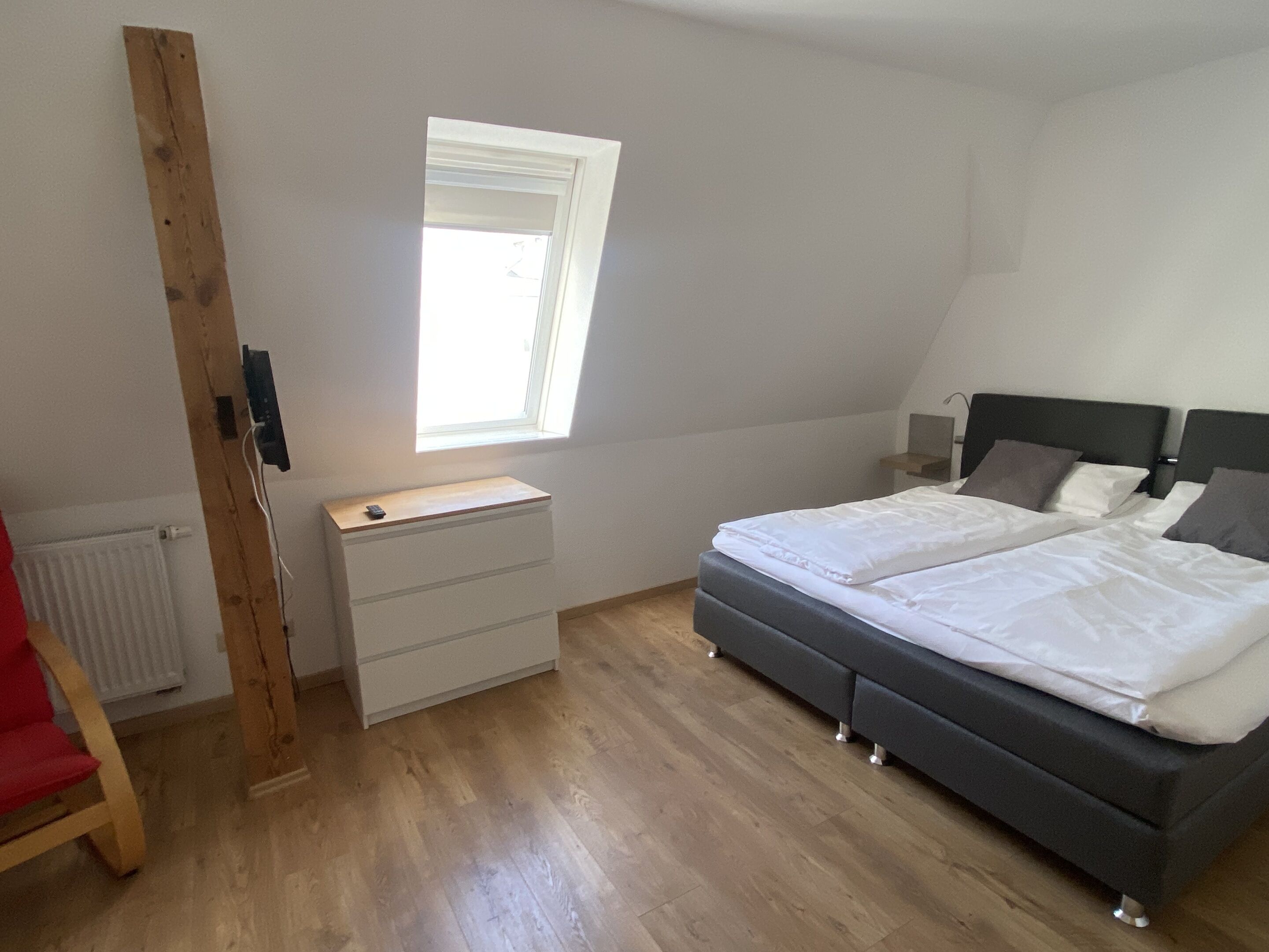 A bedroom with a bed, a bedside table, a window, a radiator, and a wooden chair.
