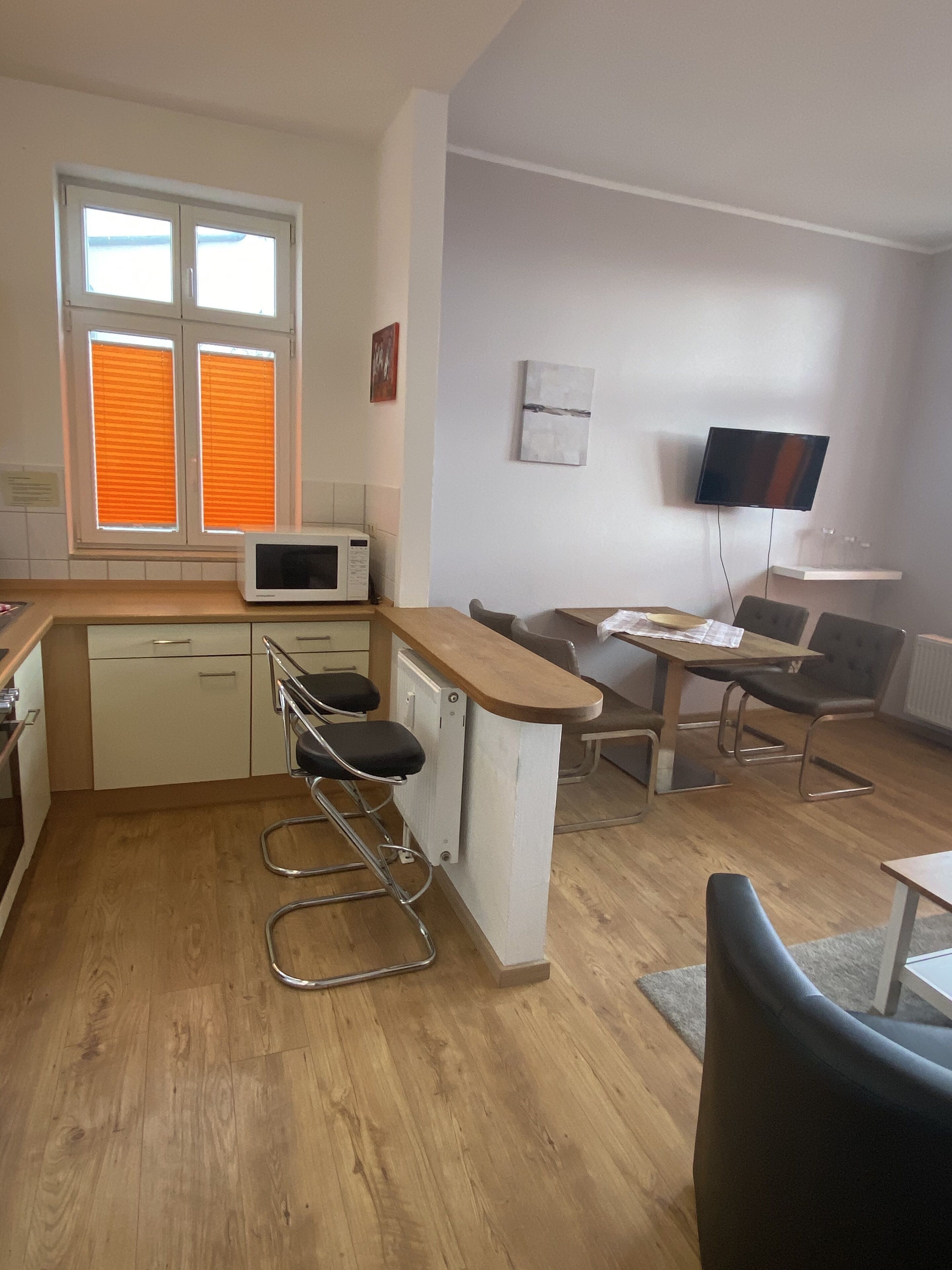 A compact kitchen with a microwave, bar stools, a dining table with chairs, a TV mounted on the wall, and a window with blinds.