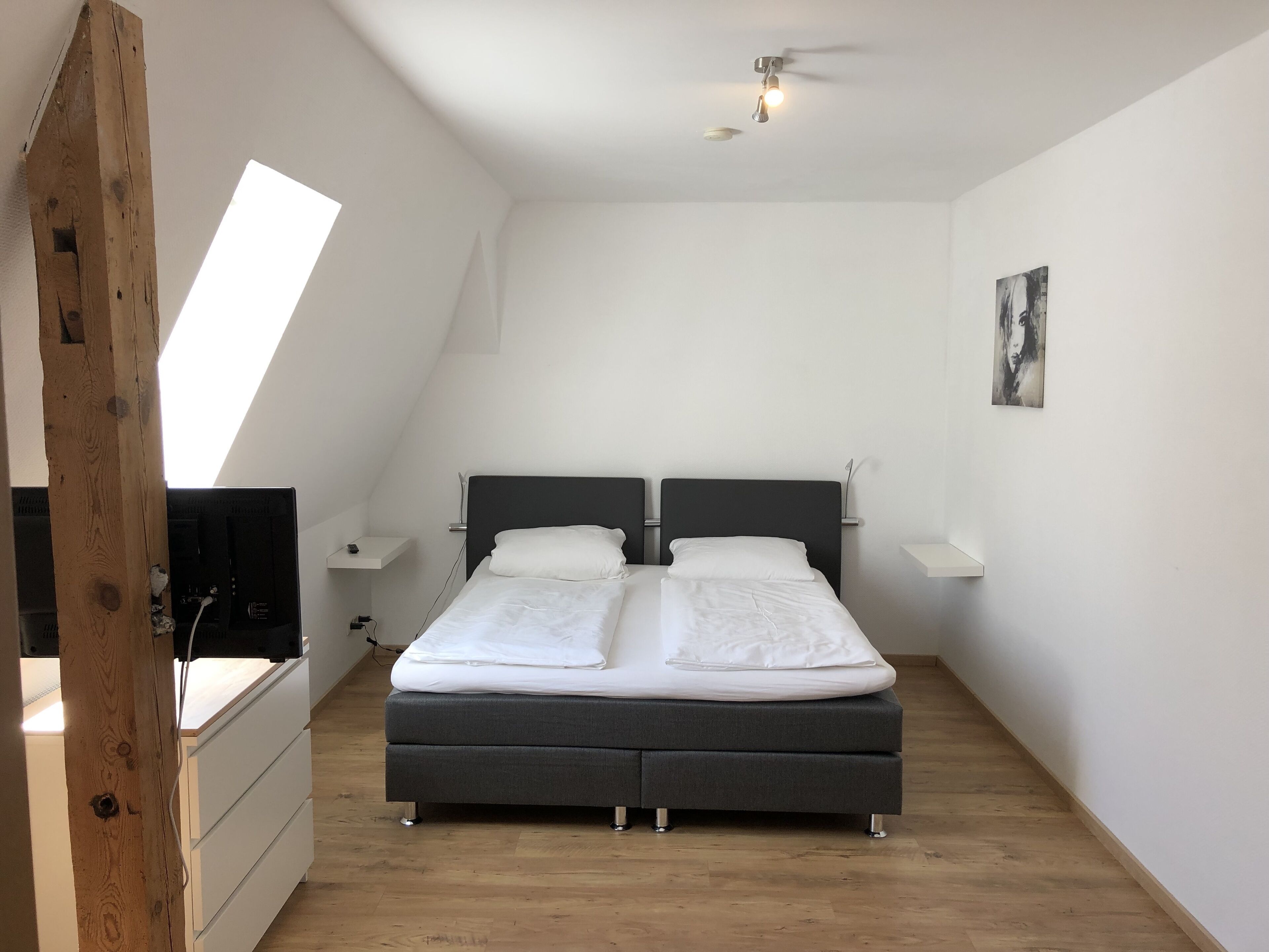 A bedroom with a bed, two bedside tables, a ceiling fan, a painting, and a wooden beam structure.