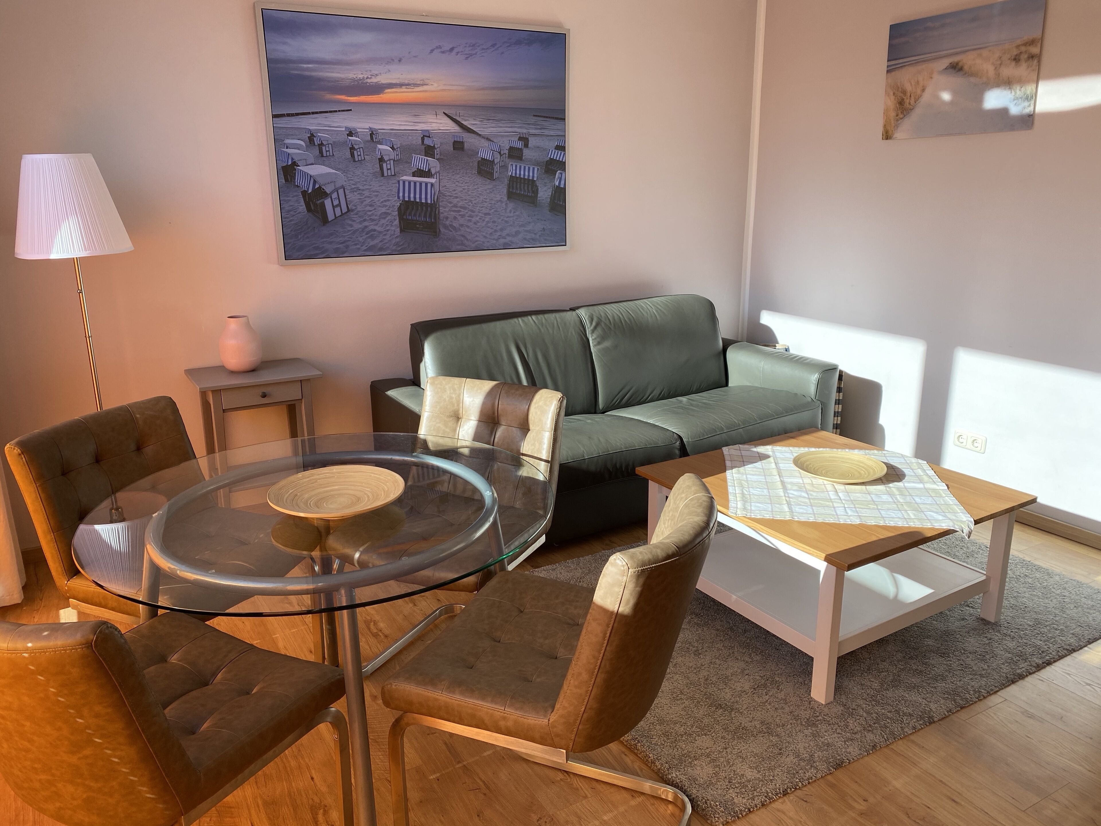 A modern living room with a glass dining table, leather sofas, and a large beach-themed painting on the wall.