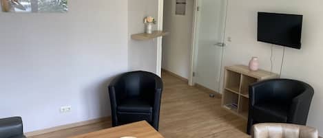 Un salon moderne avec une table basse en bois, deux chaises noires, un canapé beige, une porte en verre donnant sur une autre pièce, un téléviseur fixé au mur et une petite étagère avec un vase.