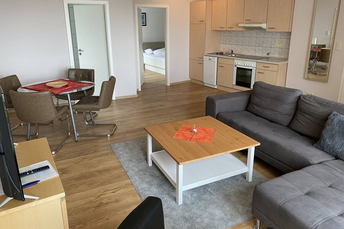 Un salon moderne avec un canapé gris, une table basse en bois et un coin repas avec une nappe rouge. On aperçoit au fond une cuisine avec des meubles en bois et une cuisinière.