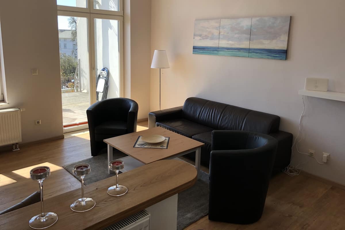 Un salon moderne avec un canapé en cuir noir, une table basse en bois et deux fauteuils noirs. Un tableau représentant une scène de plage est accroché au mur, et une lampe est visible.