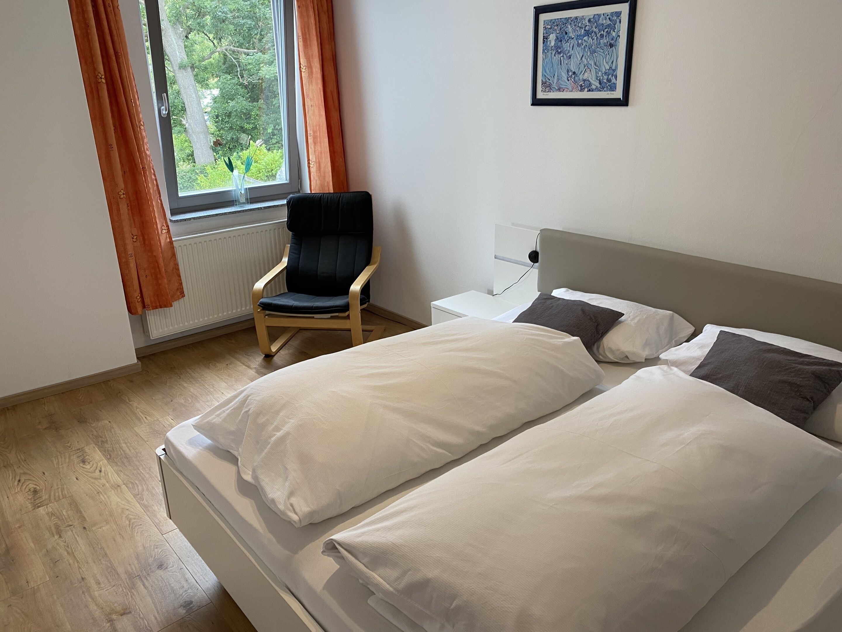 Une chambre à coucher avec un lit, une chaise, une table de chevet, une fenêtre avec des rideaux et un tableau encadré au mur.