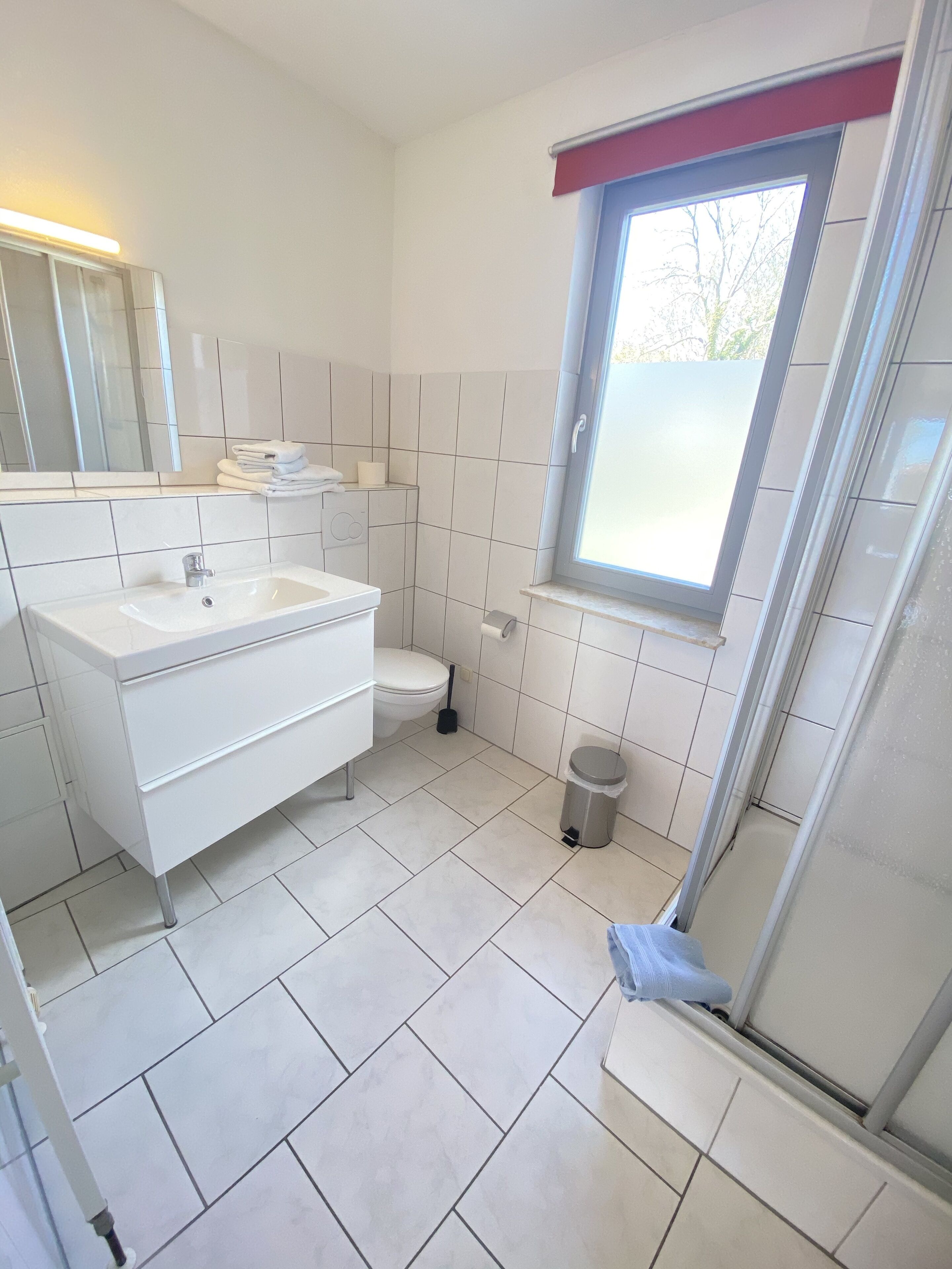 A modern bathroom with a white sink, toilet, and shower. There is a window with a red frame, and the floor is tiled.
