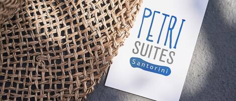 A white sign with the text "PETRI SUITES Santorini" placed on a concrete surface next to a woven basket.
