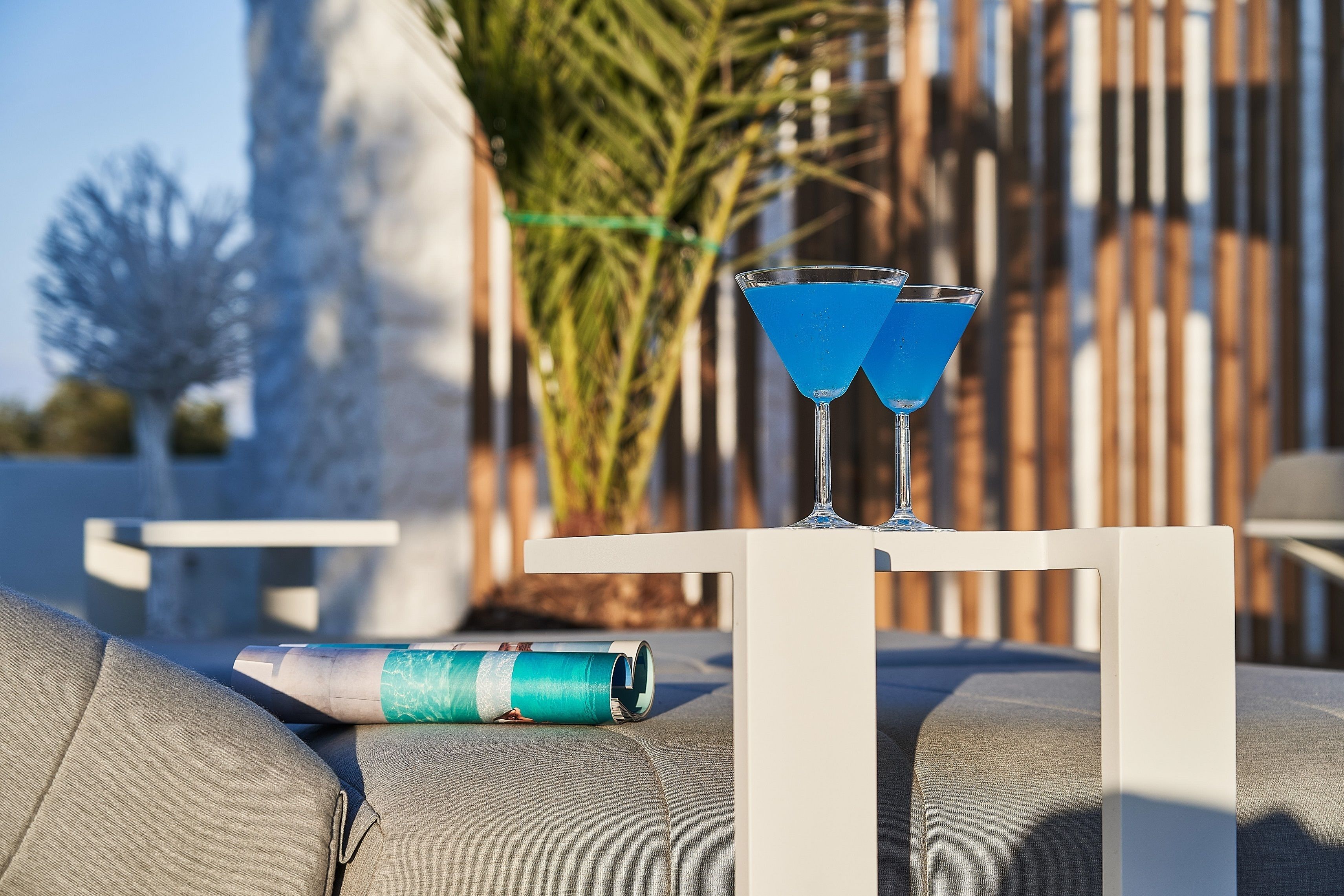 A table with two blue drinks and a rolled-up magazine on a grey couch.