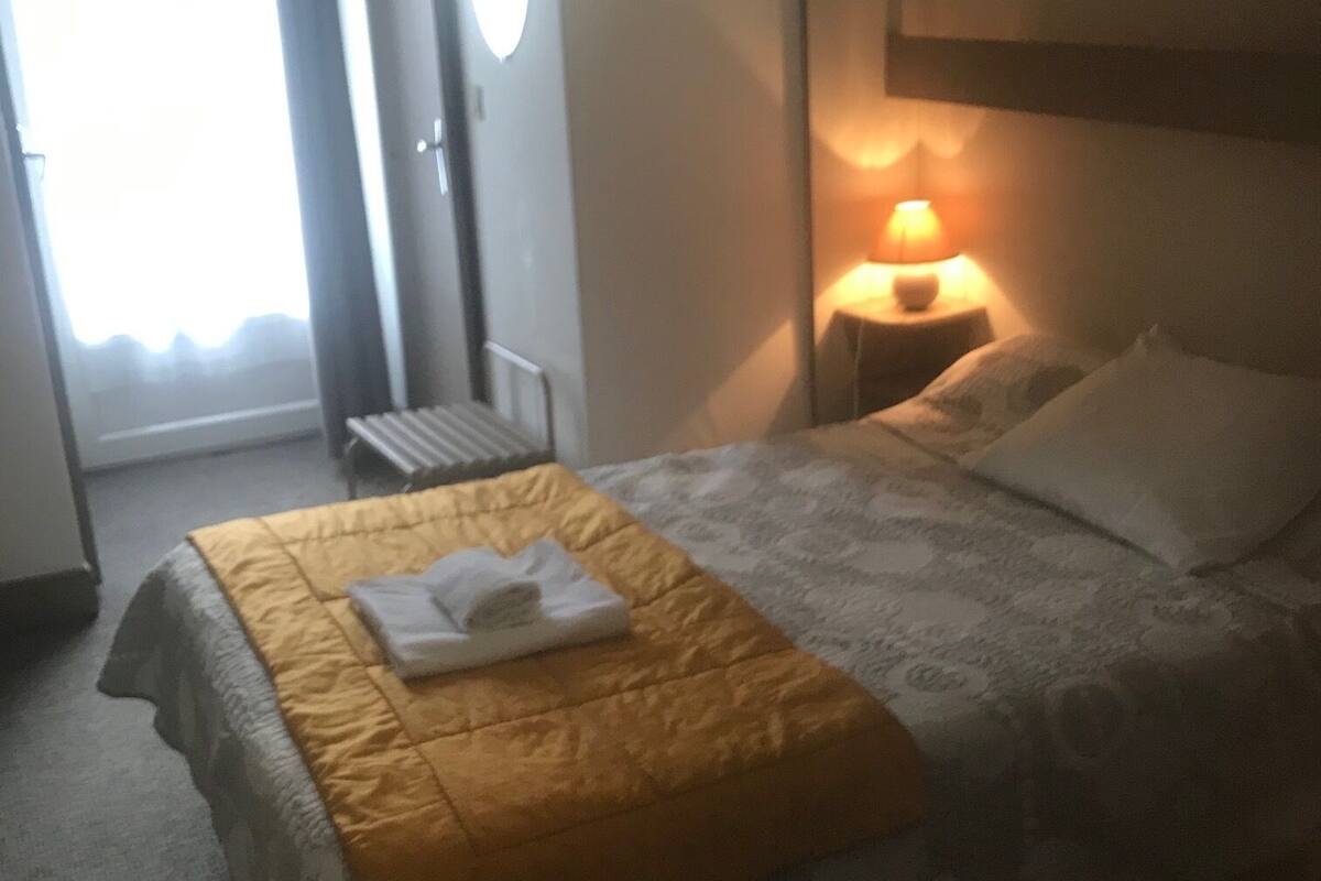 A hotel room with a bed, a nightstand with a lamp, a window with curtains, and a radiator.