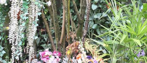 Un jardín con diversas plantas y flores, una valla metálica y un pájaro posado en una rama.