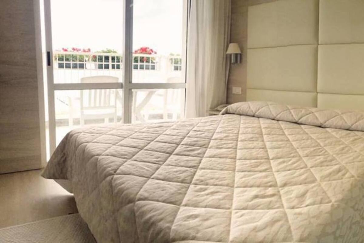 A hotel room with a large bed, a small bedside table, a lamp, and a view of the outside.