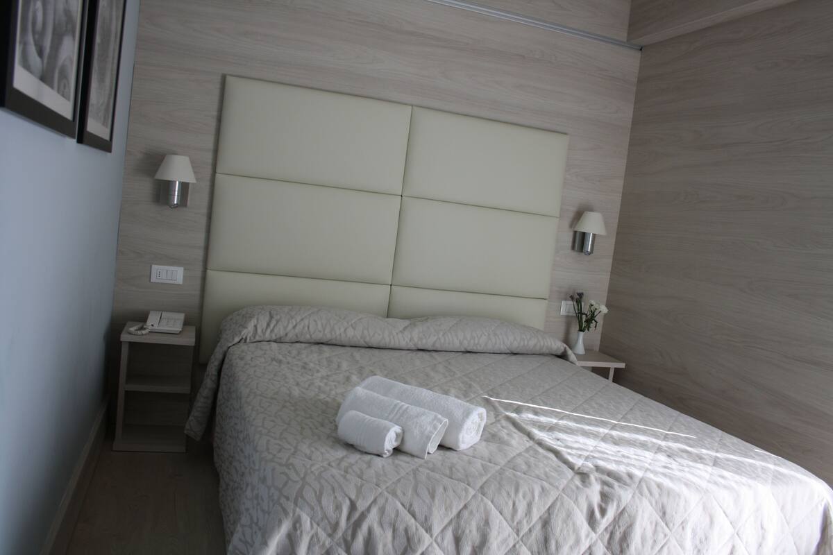 A neatly made bed with a quilted headboard, two bedside lamps, a nightstand with a phone, and a vase with flowers.