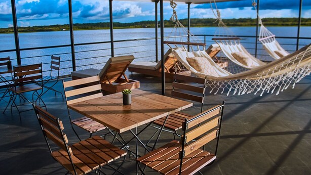 A covered outdoor seating area with wooden tables and chairs, a hammock, and a view of a lake. - Jungle Experiences La Perla (Nauta)