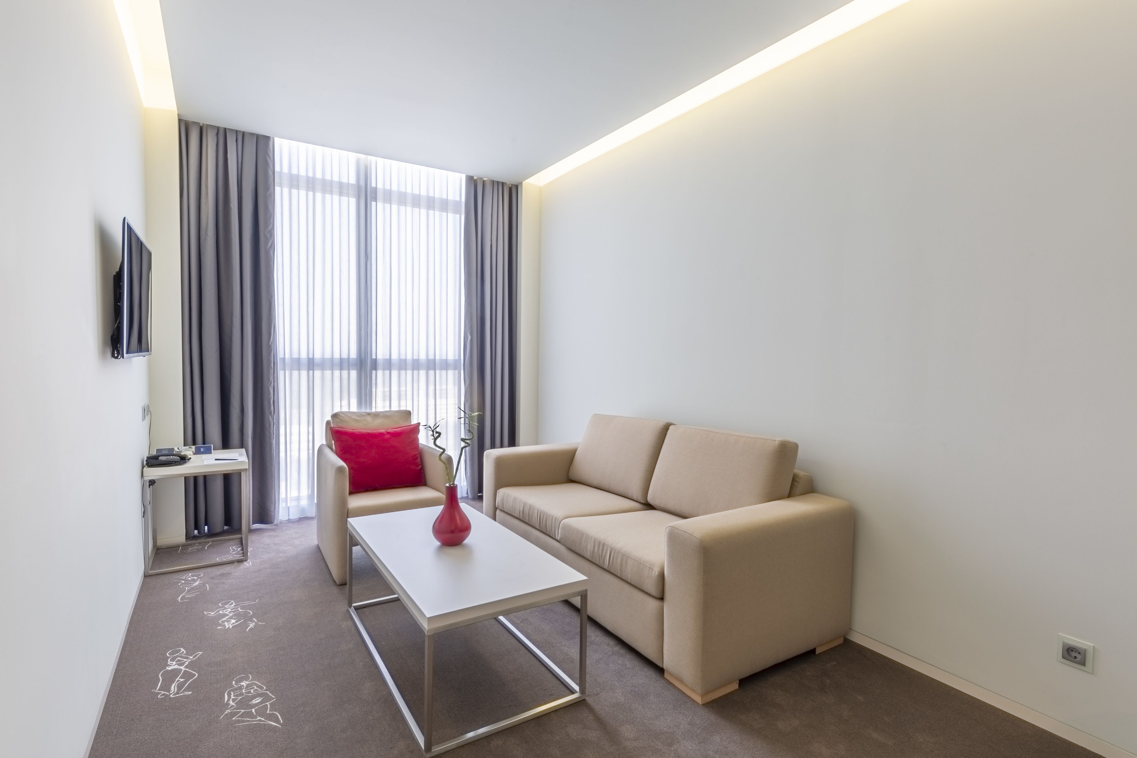 Una sala de estar moderna con un sofá, una mesa de centro, un televisor montado en la pared y un ventanal con cortinas.